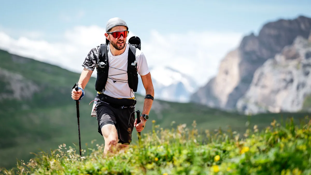 Luciano Fochi in salita con i bastoncini, sui prati di montagna sotto le pareti rocciose
